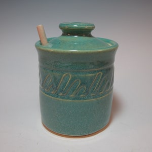 May include: A teal ceramic honey pot with a wooden dipper. The pot has a decorative design around the middle.