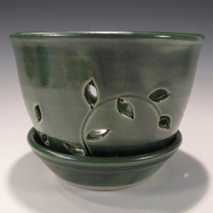 May include: A dark green ceramic planter with leaf-shaped cutouts and a matching saucer. The planter has a glossy finish and a rounded shape, suitable for small plants or succulents. The design features a decorative leaf pattern.