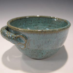 May include: A handmade ceramic bowl with a teal glaze and a textured handle. The bowl has a rounded shape and a speckled finish, with a darker rim. The handle is intricately designed, adding a unique touch to this functional art piece.