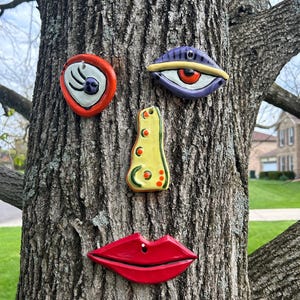 May include: A whimsical tree face sculpture composed of ceramic pieces. The face includes a red and white eye, a purple and yellow eye, a yellow nose, and red lips with a piercing. The pieces are attached to a tree trunk.