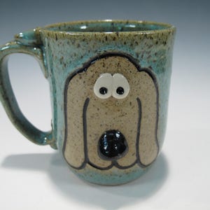 May include: A ceramic mug with a dog face design. The mug has a turquoise exterior with a tan dog face, black nose, and white eyes. The handle is turquoise. The mug is approximately 4 inches tall.