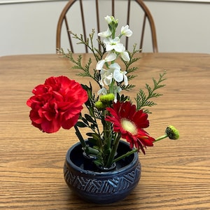 May include: A small, dark blue ceramic vase with a textured pattern holds a floral arrangement. The arrangement includes a large red carnation, a red gerbera daisy, white stock flowers, and green foliage. The vase sits on a wooden table.