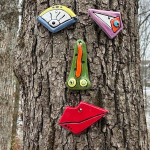 May include: A collection of whimsical ceramic face elements, including a yellow and blue fan shape, a pink triangle with an eye, a green nose, and red lips. The pieces are attached to a tree trunk.