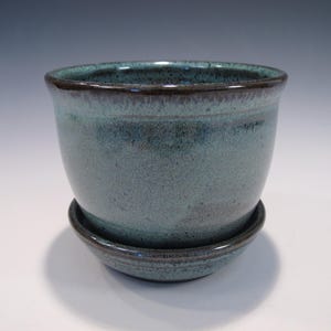 May include: A handcrafted ceramic planter in a teal glaze with a dark brown rim. The planter sits on a matching saucer. The pottery has a speckled texture and a glossy finish, suitable for indoor or outdoor use.