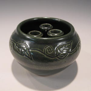 May include: A dark green ceramic bowl with three circular openings inside. The bowl has a rounded shape and is decorated with a leaf and vine pattern around the exterior. The bowl is likely used for floral arrangements.