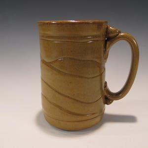 May include: A handmade, tan-colored ceramic mug with a large handle. The mug features a cylindrical shape with horizontal, wave-like grooves around the body. The mug is suitable for coffee, tea, or other beverages.