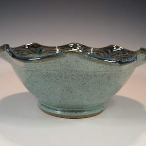 May include: A handmade ceramic bowl with a speckled, light teal glaze. The rim has a wavy design with a dark blue and brown accent. The bowl sits on a small, round base.
