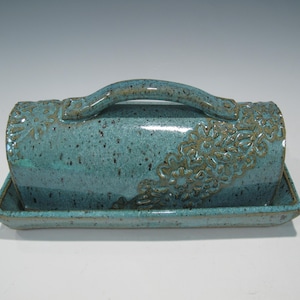 May include: A turquoise ceramic butter dish with a matching lid and handle. The lid and base are rectangular, with a floral design on the lid. The dish has a speckled appearance.