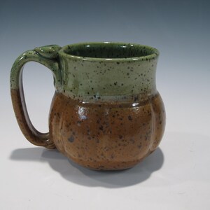 May include: A ceramic mug with a rounded, ribbed body featuring a two-tone glaze. The upper portion is a light sage green with dark speckles, while the lower section and handle are a warm, earthy brown with darker brown speckles throughout.