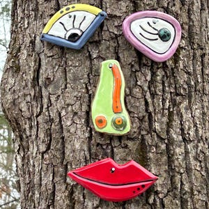 May include: A whimsical face sculpture made of ceramic pieces attached to a tree trunk. The face features a blue and yellow eye, a pink eye, a green nose, and red lips with a black stud. The artwork is colorful and abstract.