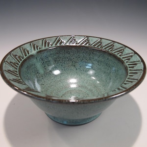 May include: A ceramic bowl with a speckled teal glaze and a dark brown rim. The rim features a repeating geometric pattern. The bowl's interior reflects light, and the overall design is simple yet elegant.