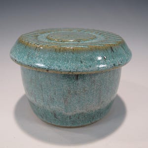 May include: A turquoise ceramic butter dish with a lid. The lid has a textured, concentric circle design. The dish has a rounded shape with a slightly speckled appearance. The rim of the lid has a brown accent.
