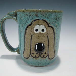 May include: A handmade ceramic mug with a teal glaze and a whimsical dog face design. The dog's face is tan with black accents, white eyes, and a black nose. The mug has a large handle and is suitable for hot or cold beverages.
