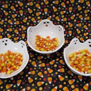May include: Three white ceramic bowls shaped like ghosts, each featuring two small circular cutouts for eyes and a larger cutout for a mouth. Each bowl is filled with orange, yellow, and white candy corn pieces, arranged on a black fabric background.