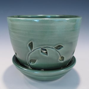 May include: A teal ceramic planter with a matching saucer. The planter features leaf-shaped cutouts arranged in a decorative pattern. The glazed finish gives the pot a smooth, glossy appearance. This handcrafted pot is suitable for indoor or outdoor use.