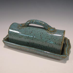 May include: A turquoise ceramic butter dish with a handle. The dish has a rectangular base with a curved edge and a matching lid. The glaze has a speckled appearance with brown accents.