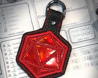 May include: A red, iridescent D20 dice keychain with black trim and a black leather strap. The keychain is on a tabletop with a Dungeons & Dragons character sheet, dice, and a Dungeon Supplies sticker.