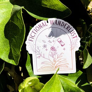 May include: A white sticker with the words "FICTIONAL WANDERLUST" in a circular design. The sticker features an open book with flowers and a crescent moon. The sticker is surrounded by green leaves.