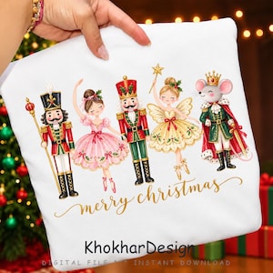 May include: A white t-shirt featuring a watercolour illustration of a nutcracker, a ballerina in a pink dress, a second nutcracker, a fairy with a star wand, and a mouse king in a crown. The text "merry christmas" and "KhokharDesign" appear.