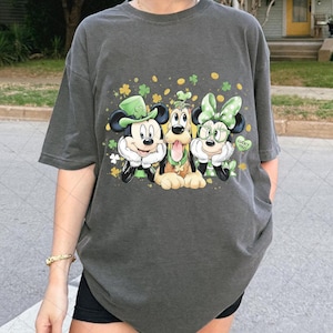 Puede incluir: Camiseta gris oscuro con Mickey Mouse, Pluto y Minnie Mouse vestidos para el Día de San Patricio. Mickey lleva un sombrero verde, Pluto un collar verde y Minnie un lazo y gafas verdes. La camiseta tiene un gráfico con tréboles y la frase "Lucky You!"