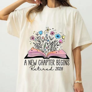 May include: Cream-colored t-shirt featuring a floral design emerging from an open book. The design includes pink, blue, and yellow flowers, with the text "A New Chapter Begins Retired 2026" in black script.