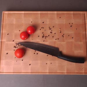 Puede incluir: Una tabla de cortar de madera con un cuchillo de mango oscuro y tres tomates rojos. Se ven granos de pimienta esparcidos. La tabla tiene un color de madera clara y un patrón de bloques cuadrados.