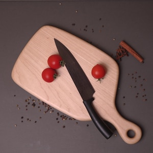 May include: A light-colored wooden cutting board with a black knife and three red cherry tomatoes. The board has a handle with a hole. Scattered peppercorns, star anise, and a cinnamon stick are also present.