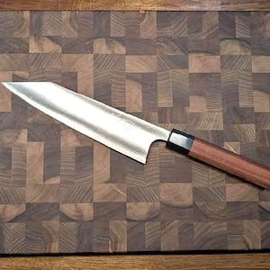 Puede incluir: Un cuchillo de chef de acero inoxidable con mango de madera descansa sobre una tabla de cortar de carnicero marrón oscuro. La tabla de cortar tiene un patrón de cuadros. La hoja del cuchillo es brillante y refleja la luz.