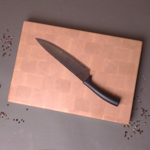 Puede incluir: Una tabla de cortar de madera clara con un patrón de cuadros y un cuchillo con mango negro en la parte superior.
