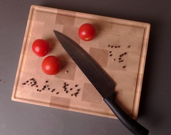 May include: A wooden chopping board with a black knife, three red tomatoes, and peppercorns. The board has a light wood colour and a square pattern. The knife is positioned diagonally across the board.