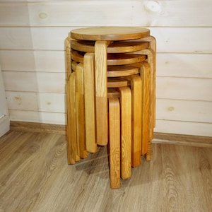 May include: A stack of wooden stools with round seats and rectangular legs. The stools are made of light-colored wood and are arranged in a vertical stack. The stools are a light brown color and are stacked on top of each other.
