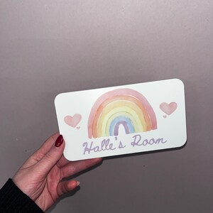 May include: A rectangular sign with a watercolor rainbow design in pastel colors. The sign has the text "Halle's Room" in a script font, with two pink hearts. The sign is held by a person.