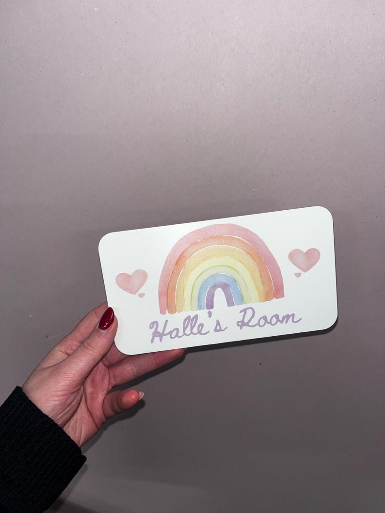 May include: A rectangular sign with a watercolor rainbow design in pastel colors. The sign has the text "Halle's Room" in a script font, with two pink hearts. The sign is held by a person.
