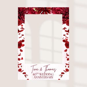 May include: A rectangular photo booth frame featuring a white centre cutout surrounded by deep red floral arrangements, including roses and dahlias, with scattered red petals. The text reads Tina & Thomas 40TH WEDDING ANNIVERSARY.