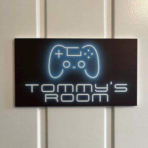 May include: A rectangular sign with a black background and a neon blue controller graphic. The text "Tommy's Room" is displayed in white, below the controller. The sign is mounted on a white door.