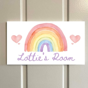May include: A rectangular sign with a watercolor rainbow design in shades of pink, orange, yellow, green, blue, and purple. Two pink hearts flank the rainbow. The text "Lottie's Room" is written in a light purple script font.
