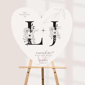 May include: A heart-shaped wedding sign in white with black lettering. It features the text THE WEDDING OF, the initials L and J decorated with black floral line art, and the names Lauren & Joe, location BILCARY HALL, and date 30.05.2026.