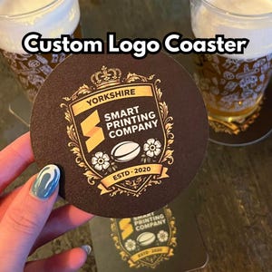 May include: A round dark brown coaster featuring a gold-coloured crest logo with the text "YORKSHIRE SMART PRINTING COMPANY ESTD 2020" and an illustration of a rugby ball and flowers. The text "Custom Logo Coaster" is overlaid at the top of the image.