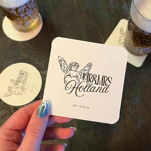 May include: A white square coaster featuring a black angel illustration and the text "MR & MRS Holland" with "EST. 18.09.20". A round coaster with the same design is also visible. The coasters are on a dark surface.