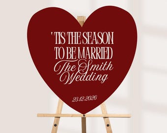 May include: A heart-shaped dark red sign displayed on a light wooden easel. The sign features white text that reads 'TIS THE SEASON TO BE MARRIED, The Smith Wedding, 23.12.2026' in a combination of bold block lettering and elegant script.