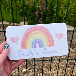 May include: A rectangular white sign with a watercolor rainbow design and two pink hearts. The text "Emily's Room" is written in light blue script. The sign is held in front of a green fence and foliage.