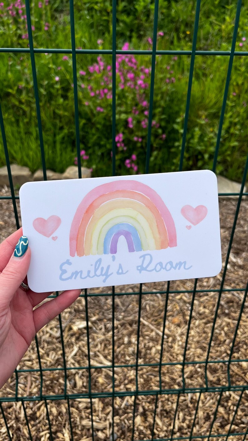 May include: A rectangular white sign with a watercolor rainbow design and two pink hearts. The text "Emily's Room" is written in light blue script. The sign is held in front of a green fence and foliage.