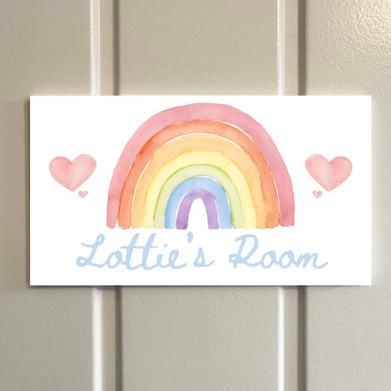 May include: A rectangular sign with a watercolor rainbow design in shades of pink, orange, yellow, green, blue, and purple. Two pink hearts flank the rainbow. The text "Lottie's Room" is written in light blue script.