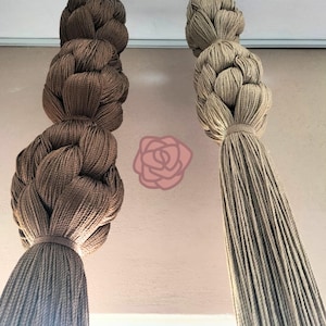 May include: Two decorative macrame tassels hanging from the ceiling. One is a dark brown color, and the other is a light beige. The tassels are made of braided rope and are tied at the top with a matching cord.