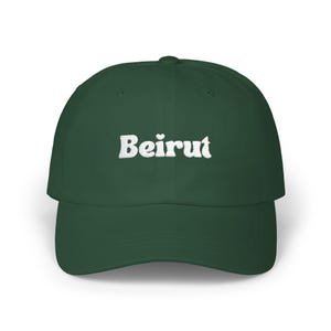 May include: A dark green baseball cap with the word "Beirut" in white, stylized text across the front. The cap has a curved brim and a rounded crown. The text includes a small heart above the "i".