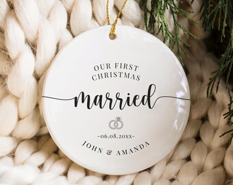 May include: A white ceramic Christmas ornament with the text "OUR FIRST CHRISTMAS married" in black script. Below the text are two wedding rings and the date "-06.08.20xx-" and the names "JOHN & AMANDA". The ornament hangs from a gold string.
