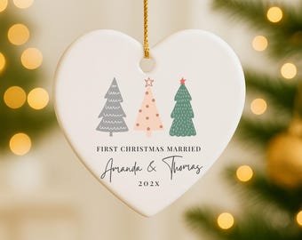 May include: Heart-shaped ceramic Christmas ornament with three illustrated Christmas trees in gray, pink, and green. The text "FIRST CHRISTMAS MARRIED" and names "Amanda & Thomas" are printed below the trees, with the year "202X" below the names.