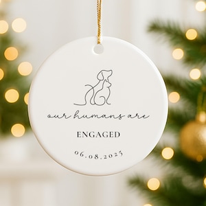 May include: A white ceramic ornament with a gold string for hanging. The ornament features a line drawing of a dog and cat, with the text "our humans are ENGAGED 06.08.2025". The ornament is round and has a glossy finish.