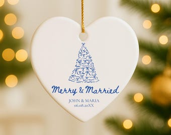 May include: Heart-shaped white ceramic Christmas ornament with a blue Christmas tree design. The ornament has the text "Merry & Married" and the names "JOHN & MARIA" with a date. A gold string is attached for hanging.