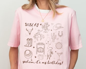 May include: Pink t-shirt with a western-themed graphic. The design includes a bull skull, cowboy boots, a horseshoe, a cactus, and the text "Wild 40" and "yeehaw, it's my birthday!"